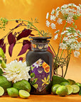 An elegant dark glass bottle labeled "Romance in Ravello" from Magic Hour is surrounded by fresh green limes, lime wedges, and assorted flowers against a vibrant yellow-orange background. This visually rich composition evokes the natural beauty and freshness of the Amalfi Coast, enhanced by hints of chocolate, passion fruit, and rose cream.
