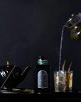 A hand pours **Magic Hour Renewal: Peach-Goji-Rose Oolong Tea** from a glass kettle into a glass filled with ice beside a black teapot. Behind the glass, an ornate bottle is displayed against a dark background. Two more glasses with golden straws rest nearby, creating an elegant drink setup.