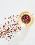 A glass tea infuser filled with loose leaf tea from the Classics - Luxe Sampler Sips collection, showcasing visible flower petals and herbs, sits on a white surface. Some of the colorful tea leaves and petals are scattered around the infuser, creating a vibrant visual contrast with the background.
