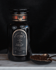 A dark glass jar labeled "Club Magic Hour" rests on a marble surface against a black background. Beside the jar, a brass spoon lies on a small dish filled with loose tea leaves, including Rooibos. The label showcases an hourglass along with the words "Time for Infinity" and "Hand-Crafted Tea.