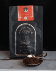 A package of "Rise & Shine Herbal Tea Case Packs" from Club Magic Hour sits on a marble surface. The black bag, adorned with gold and red detailing, is accompanied by a spoonful of rooibos tea leaves in front. The design features a celestial theme and reads "Time for Infinity.