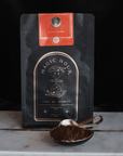 A black, resealable bag labeled "Rise & Shine Black Tea" with a celestial design and the phrase "Time for Infinity" is displayed upright. A red label at the top says "Club Magic Hour." In front, a spoon rests in a small bowl filled with organic loose leaf tea.