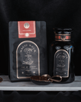 A photograph of two Club Magic Hour tea products on a dark background. To the left is a black and red "Rise & Shine Herbal Tea Case Packs" pouch with a celestial design. To the right, there's a matching black tea jar. A golden spoon with chaga mushroom-infused tea leaves is placed in front of the jar.