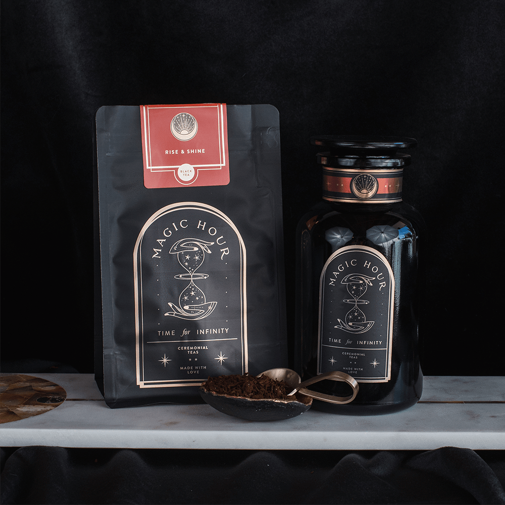 A photo of two Club Magic Hour products. On the left is a flat pouch labeled "Rise & Shine Black Tea," featuring organic loose leaf tea. On the right is a black jar labeled "Time & Infinity," with both items adorned with celestial-themed designs and a teaspoon filled with tea leaves placed in front.