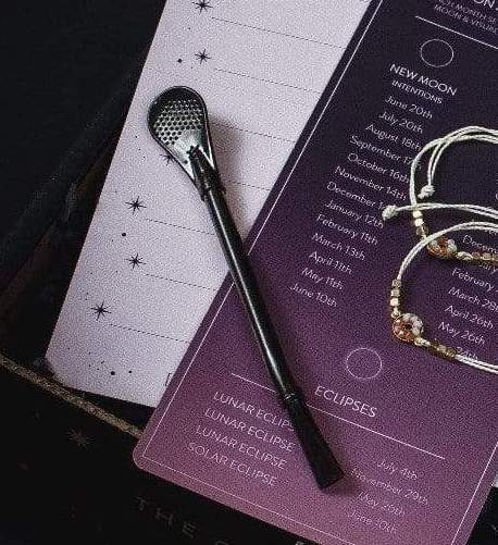 A close-up of a planner page listing lunar events and new moon intentions, with a black pen and a beaded bracelet resting on top. The planner, perfect for jotting down thoughts while sipping Magic Hour's Moon Lover: Herbal Immunitea Ritual Gift, has a dark and light gradient design transitioning from purple to white, adorned with small star illustrations.