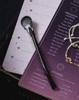 A close-up of a planner page listing lunar events and new moon intentions, with a black pen and a beaded bracelet resting on top. The planner, perfect for jotting down thoughts while sipping Magic Hour's Moon Lover: Herbal Immunitea Ritual Gift, has a dark and light gradient design transitioning from purple to white, adorned with small star illustrations.