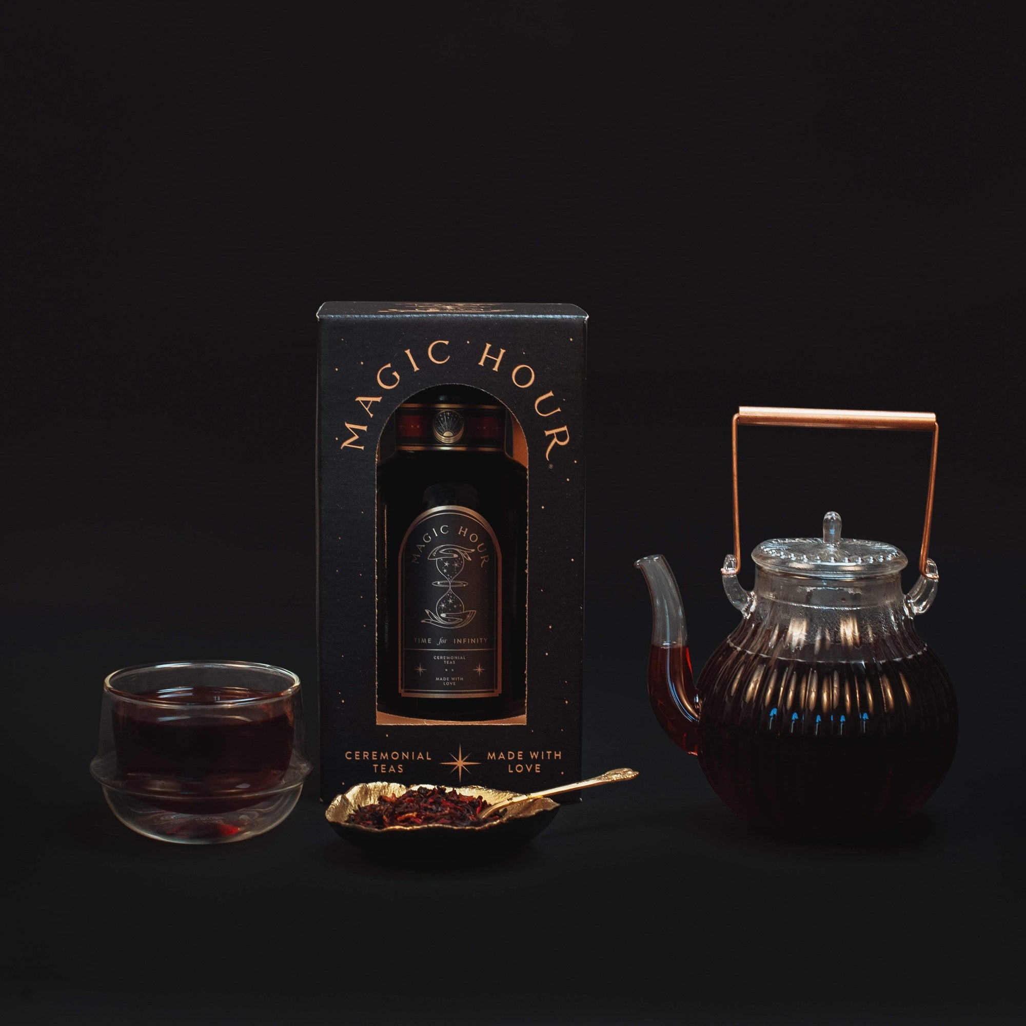 A Best Sellers of Magic Hour Box by Magic Hour in its packaging is displayed against a black background. Beside it is a transparent glass teapot filled with dark, loose leaf tea and a matching teacup. A gold spoon with loose tea leaves sits in front of the teapot and cup.