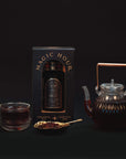 A Best Sellers of Magic Hour Box by Magic Hour in its packaging is displayed against a black background. Beside it is a transparent glass teapot filled with dark, loose leaf tea and a matching teacup. A gold spoon with loose tea leaves sits in front of the teapot and cup.
