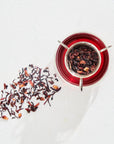A clear cup of red tea from the Classics - Luxe Sampler Sips collection is viewed from above on a white surface. Inside the cup, a metallic infuser holds Bohemian Breakfast tea leaves. Loose tea leaves are scattered to the left of the cup, casting a shadow in the sunlight.