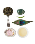A composition displaying a spoonful of Ti Quan Yin Oolong: Tea of Patient Compassion leaves, a green gemstone, a peacock feather, a fresh leaf, a polished shell with a pink rose petal inside, and a cup of light-colored tea. All arranged neatly on a white background to highlight the wellness benefits of Magic Hour Ti Quan Yin Oolong: Tea of Patient Compassion.