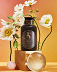 A black perfume bottle with a gold label reading "Sagittarius Tea of Good Fortune & Abundance - Magic Hour" is displayed on a gold pedestal against a gradient background of orange and pink. White flowers and two clear spheres, one pink and one iridescent, surround the bottle, evoking the elegance of Magic Hour tea rituals.