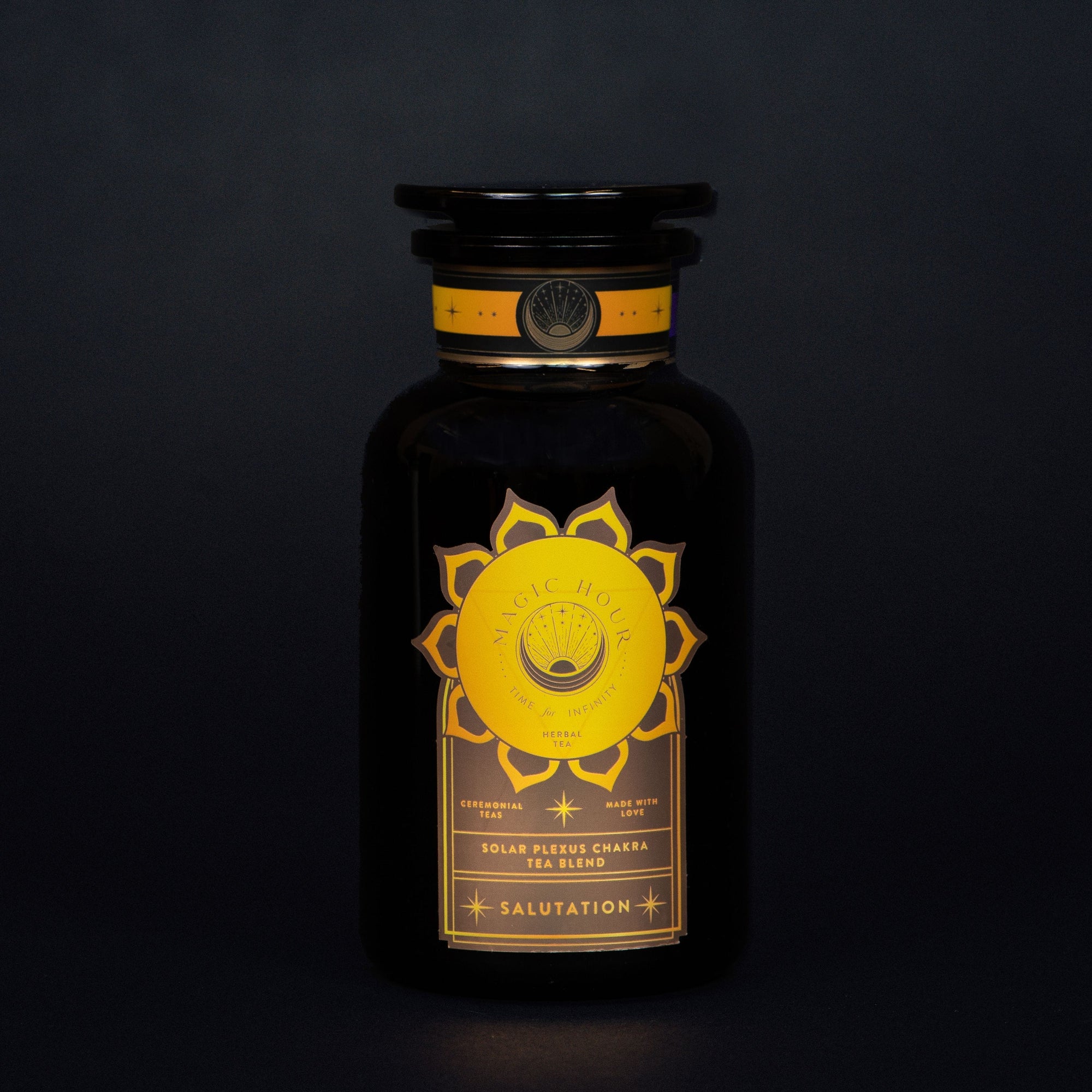 A black glass bottle with a wide black lid, labeled "Salutation™ Tea for the Solar Plexus Chakra" in a golden circle design featuring the solar plexus chakra symbol. The label states: "Solar Plexus Chakra Tea Blend," "Salutation," and "Ceremonial," on a dark background, perfect for achieving chakra balance with organic ingredients.