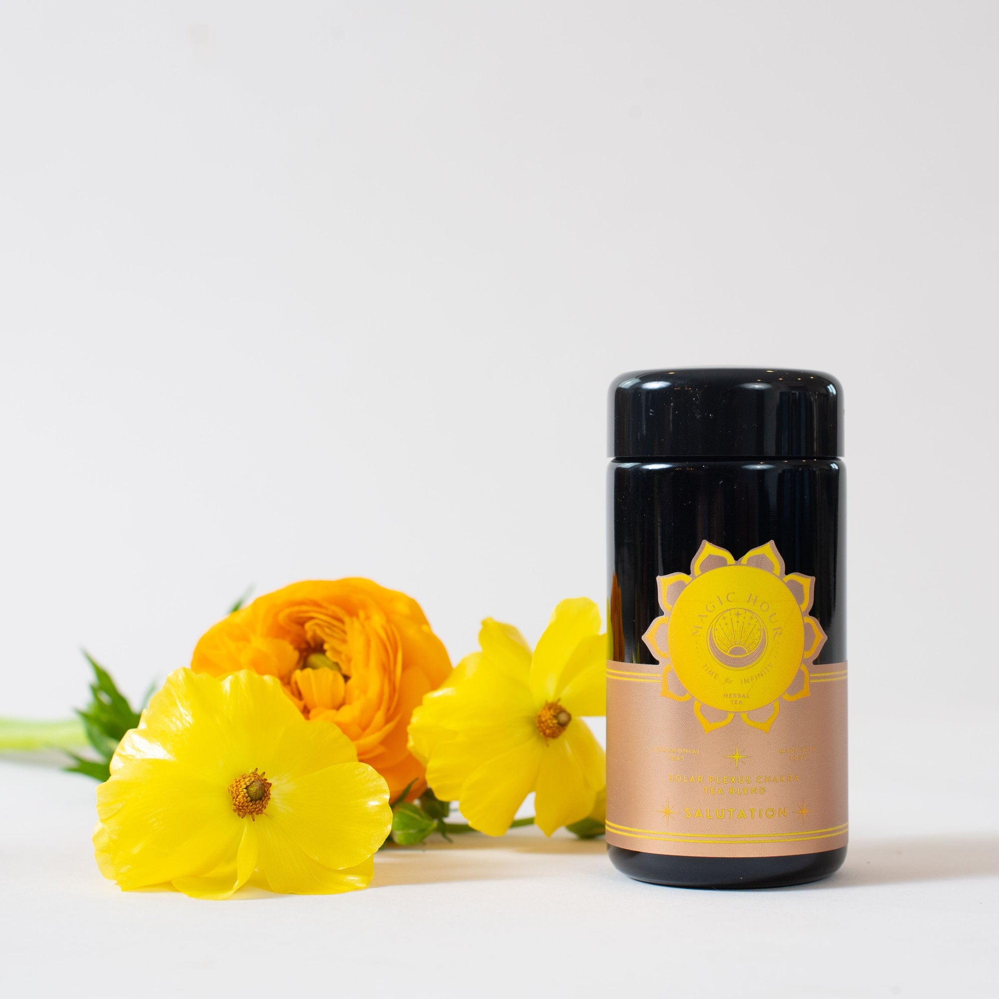A black cylindrical container with a yellow label featuring a sun-like emblem sits next to vibrant yellow and orange flowers on a white surface. The container's label reads "Salutation™ Tea for the Solar Plexus Chakra," highlighting its organic ingredients. The background is plain and light-colored.