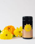 A black cylindrical container with a yellow label featuring a sun-like emblem sits next to vibrant yellow and orange flowers on a white surface. The container's label reads "Salutation™ Tea for the Solar Plexus Chakra," highlighting its organic ingredients. The background is plain and light-colored.