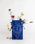 A blue geometric-patterned tea bag labeled "Sapphire - Apple-Plum-Spice White Tea Case Packs" from Magic Hour is centered against a white background. The tea bag is surrounded by an arrangement of flowers, including white roses and purple blooms, emerging from behind it.