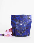 A blue, geometrically designed pouch labeled "Sapphire - Apple-Plum-Spice White Tea Case Packs" from Magic Hour is displayed against a white background. Beside the pouch, there is a light pink orchid and a small purple flower, adding a touch of natural elegance to the image of this calming grounding tea.