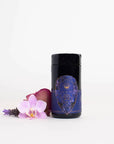 The "Sapphire - Apple-Plum-Spice White Tea Case Packs" by Magic Hour, housed in a black cylindrical container with a purple geometric label featuring gold details and text, stands next to a pink orchid flower and a red rose petal, all placed on a white surface with a white background.
