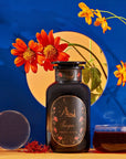 A dark jar labeled "Scorpio Tea for Sensual Brilliance" is placed on a table next to a teacup filled with Magic Hour Tea, saffron threads, and a clear orb. The background features bright orange flowers and a vibrant blue backdrop with a circular opening revealing more flowers.