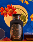 A dark jar labeled "Scorpio Tea for Sensual Brilliance" is placed on a table next to a teacup filled with Magic Hour Tea, saffron threads, and a clear orb. The background features bright orange flowers and a vibrant blue backdrop with a circular opening revealing more flowers.
