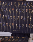 A person holds up a dark blue cloth decorated with rows of flowers in various colors. Below the flowers, the text "MAGIC HOUR" is visible. The background shows a kitchen countertop with a large black container of loose leaf tea and other tea-related items, including the Tea Ceremony Towels Bundle from Magic Hour.