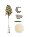 A flat lay image features a silver spoon filled with dried herbs, a beaded crescent moon, a small piece of amethyst crystal, a green leaf blade, and a white bowl containing light-colored Silver Moon White Tea from Club Magic Hour, all artfully arranged on a white background.