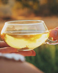 A person holds a transparent glass cup filled with light-colored Club Magic Hour Silver Moon White Tea with both hands. The outdoors background appears blurred, suggesting a warm and sunny environment, possibly during sunset. The person has a visible ring on their finger.