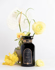A black bottle labeled "Sin Eraser™ : Puerh Tea" by Magic Hour is surrounded by various flowers, including a white anthurium and yellow ranunculus, against a white background. The bottle has a gold-accented neck and the flowers visually frame it, adding an elegant touch.