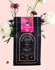 A black package of Magic Hour Symbeeosis: Beautifying Immunitea for the Queen Bee stands upright against a white background. The package is adorned with a pink label and surrounded by delicate flowers, including white ranunculus and purple blooms, arranged artfully to enhance the presentation.