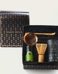 A gift box with a black and gold starry design featuring a bottle of organic tea, a matcha whisk, a gold tea strainer, and a box labeled "Ceremonial Matcha Traveler Gift Box" containing loose leaf tea. The box lid with the "Magic Hour" logo is partially removed and placed beside it.