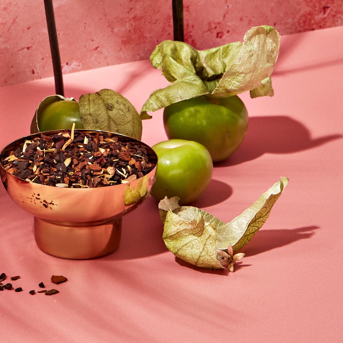 A copper bowl filled with a dark, chopped substance sits on a pink surface. Surrounding the bowl are three green tomatillos, their husks partially peeled back, all set against a pink textured background. This arrangement looks like an enticing display from the Spice Lovers Starter Set, evoking an array of exotic spice teas waiting to be discovered.