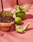 A copper bowl filled with a dark, chopped substance sits on a pink surface. Surrounding the bowl are three green tomatillos, their husks partially peeled back, all set against a pink textured background. This arrangement looks like an enticing display from the Spice Lovers Starter Set, evoking an array of exotic spice teas waiting to be discovered.