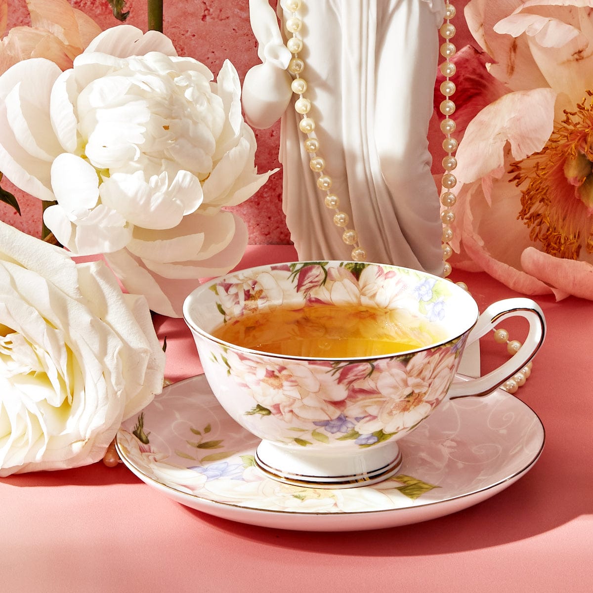 A floral-patterned teacup filled with tea sits on a matching saucer, surrounded by white peonies and peach-colored roses on a pink surface. Nearby, a Dessert Lovers Starter Set and chocolate-dipped berries add an elegant and delicate touch to the setting, complemented by the strand of pearls and white statue in the background.