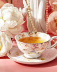 A floral-patterned teacup filled with tea sits on a matching saucer, surrounded by white peonies and peach-colored roses on a pink surface. Nearby, a Dessert Lovers Starter Set and chocolate-dipped berries add an elegant and delicate touch to the setting, complemented by the strand of pearls and white statue in the background.