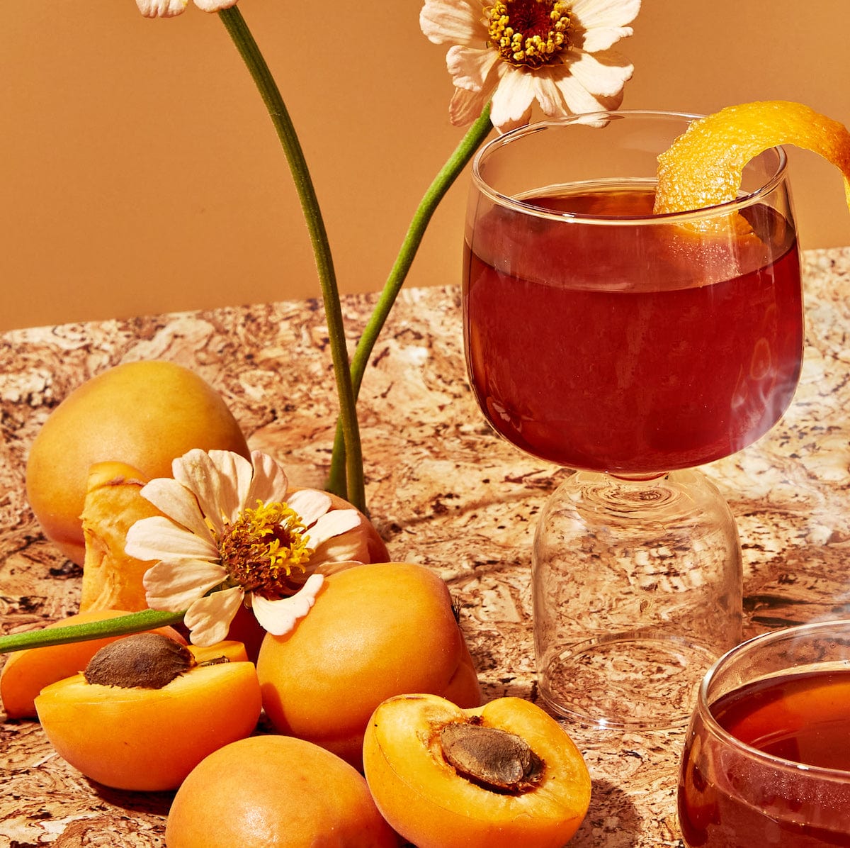 A glass of amber-colored beverage garnished with an orange peel from the Flower & Fruit Lovers Starter Set sits on a marble surface surrounded by whole apricots, apricot halves, and white flowers. Another glass from the same set, possibly filled with gemstone-wellness-tea, is partly visible in the foreground. The background exudes warm tones.