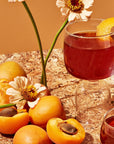 A glass of amber-colored beverage garnished with an orange peel from the Flower & Fruit Lovers Starter Set sits on a marble surface surrounded by whole apricots, apricot halves, and white flowers. Another glass from the same set, possibly filled with gemstone-wellness-tea, is partly visible in the foreground. The background exudes warm tones.