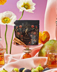 A vibrant still life featuring a black floral-embellished Magic Hour Taurus: Tea of Venusian Garden Delights package on a pink draped fabric, surrounded by tropical fruits like pineapples and mangoes, colorful flowers, two glass cups filled with organic tea, cinnamon sticks, and a sunset gradient background.