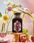 A bottle of "Breakfast in Paris-Oolong Black Tea Scented with Fruits & Flowers of Secret Parisian Gardens" is elegantly showcased on a gold stand, accompanied by flowers, fruits, and a teacup with a saucer. The background captures the essence of secret Parisian gardens, featuring a pastel gradient of soft pink, blue, and yellow tones.