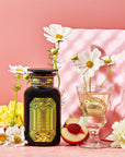 A bottle of Magic Hour's Peridot: Green Guayusa Oolong Mango-Peach Tea stands next to a wine glass with a flower and a golden strainer, all set against a pink background. White flowers, organic peach slices, and chunks of organic mango enhance the elegant display.