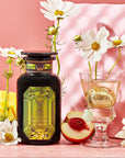 A bottle of Magic Hour's Peridot: Green Guayusa Oolong Mango-Peach Tea stands next to a wine glass with a flower and a golden strainer, all set against a pink background. White flowers, organic peach slices, and chunks of organic mango enhance the elegant display.