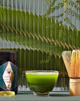 A Matcha 1111 : Highest Grade of Ceremonial Matcha kit on a green surface with a background of ferns. It includes a matcha container, a glass bowl with matcha tea, a bamboo whisk, and a small wooden spoon with matcha powder. The scene has a calm and natural ambiance, perfect for enjoying Magic Hour's organic tea selection.