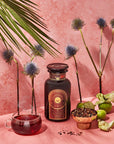 A dark bottle with a sun emblem stands on a pink surface surrounded by exotic flowers, green fruits, a teacup filled with a dark red liquid, and a bowl of organic loose leaf tea. A large green leaf dangles from above, set against a textured pink background. Magic Hour Queen of the South: Delicious Cocoa Detox Tea awaits your senses.