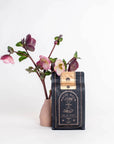 A bag of Magic Hour Ti Quan Yin Oolong: Tea of Patient Compassion stands against a white background. The bag is black with gold details. Beside it, a minimalist vase holds a stem with several purple and pink flowers. This arrangement highlights the tea's wellness benefits and is aesthetically pleasing.