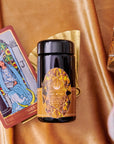 A black jar of Magic Hour’s “Topaz - Caramel Apple Oolong Tea Case Packs” with an intricate, geometric design label sits on a golden surface, perfect for cozy sips. It is accompanied by a tarot card featuring The High Priestess, a golden fan, and part of an ornate glass. The background features a rich, golden-brown fabric texture reminiscent of caramel apple crisp.