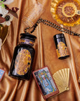 An elegant display featuring tarot cards, a black and gold bottle, glasses, a fan, and a floral arrangement on a silky, golden fabric. The tarot cards are arranged around the bottle, lending a mystical and luxurious ambiance to the setting—perfect for cozy sips of Magic Hour's Topaz - Caramel Apple Oolong Tea from the case packs.