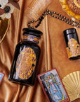 An elegant display featuring tarot cards, a black and gold bottle, glasses, a fan, and a floral arrangement on a silky, golden fabric. The tarot cards are arranged around the bottle, lending a mystical and luxurious ambiance to the setting—perfect for cozy sips of Magic Hour's Topaz - Caramel Apple Oolong Tea from the case packs.