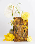 Placed in front of a white background is an elegant case pack of Magic Hour's Topaz - Caramel Apple Oolong Tea. Surrounding the case pack are vibrant yellow flowers and pristine white anthuriums, adding a touch of sophistication and color to the scene, perfect for cozy sips that warm your soul.