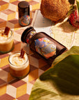 Two cocktails garnished with cinnamon sticks are placed on a patterned tile surface. Next to them is a dark glass bottle of Magic Hour's Tulum Horchata Chai Case Packs, featuring a colorful label and accompanied by a small container, hinting at an adaptogenic wellness potion. The setting includes a coconut, flower, and map partially covered by large green leaves.