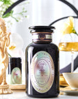 A decorative setup featuring a large dark-colored jar labeled "Pearl Tea of Beauty, Clarity, and a Sense of Peace" from Magic Hour surrounded by luxurious items, including a small bottle, a cup and saucer ready for loose leaf tea, and a delicate statue. Soft lighting and flowers create an elegant, serene atmosphere.