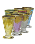 Introducing the Romantic Tea Set - Six Glasses & Pitcher: a set of six tall, footed drinking glasses in a range of colors including yellow, purple, clear, and blue. Each glass is elegantly adorned with ornate gold decorative patterns around the top and intricate designs below, capturing the classic Italian design and timeless elegance of a romantic tea experience.