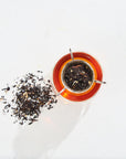A glass teacup filled with the amber-hued Mango-Ginger-Cream White Tea from the Astrology Bundle: Cancer, Leo, Virgo is placed on a white surface. The teacup features a metal tea infuser containing loose tea leaves. To the left of the teacup, there is a small pile of loose tea leaves scattered on the surface.